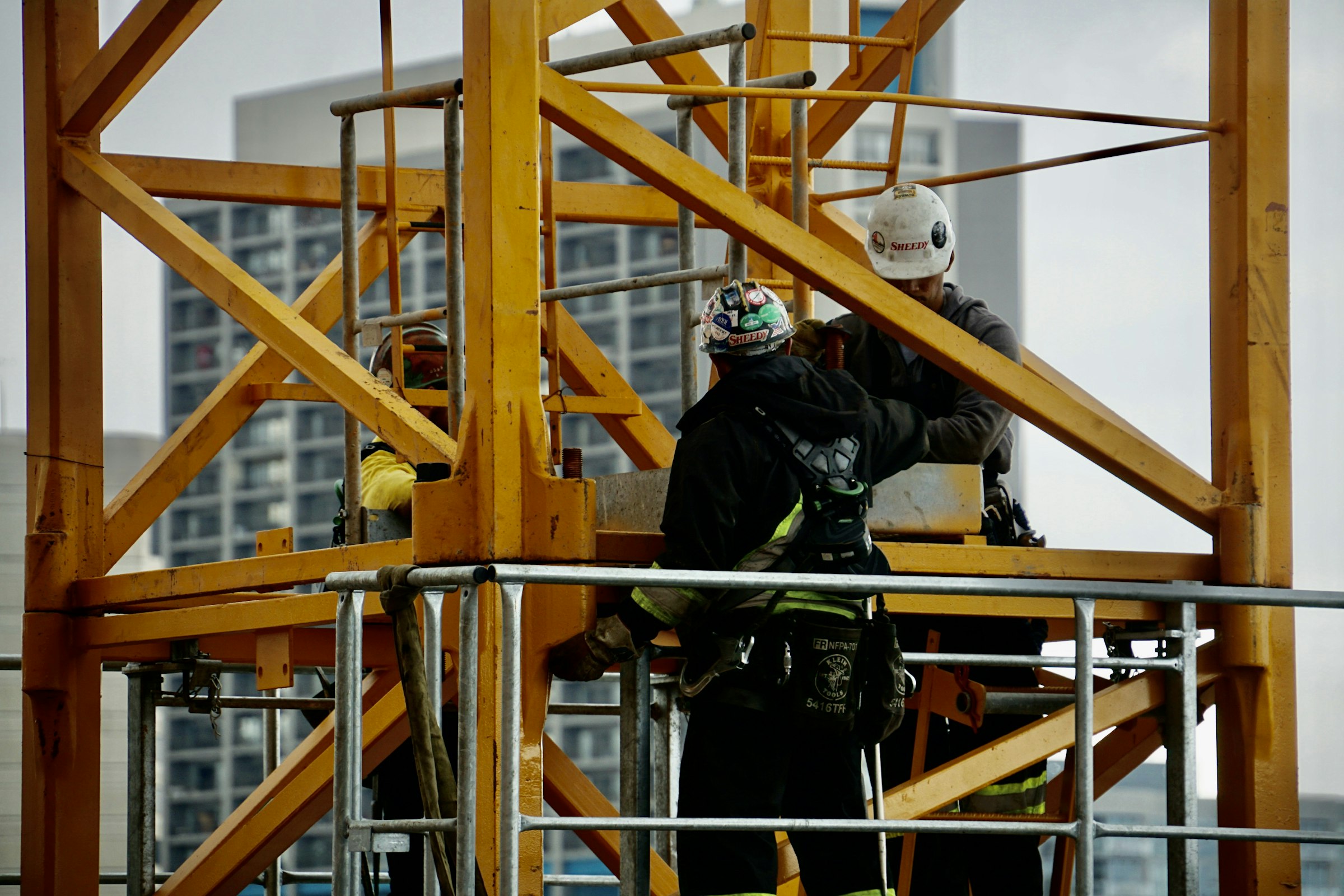 Montadores com capacete e cinto de segurança trabalhando no alto da torre de uma grua